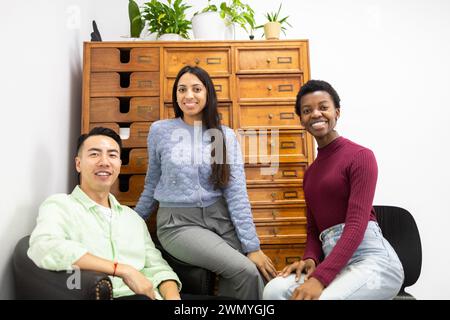 Three friends of diverse ethnicities share a moment, sitting together with warm smiles in a room decorated with plants and a vintage drawer cabinet. Stock Photo