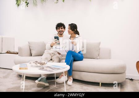 A cheerful young couple cozy up on the couch with a smartphone, sharing a moment of digital connection in a modern living room setting. Stock Photo