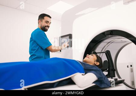 Smiling male radiologist operating a computer while preparing a female patient for a computed tomography scan in a modern medical facility. Stock Photo