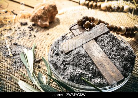 Detail of glass container with ashes and wooden cross on burlap cloth with olive leaves on a wooden table. Elevated view. Stock Photo