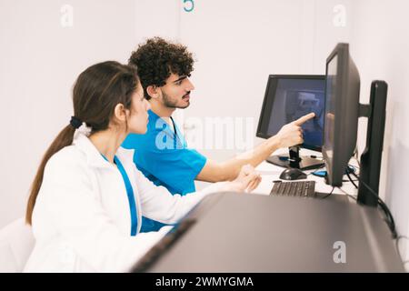 Two healthcare workers in blue scrubs are focused on a computer screen, discussing medical data in a bright office setting. Stock Photo