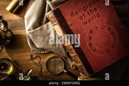 Old and deteriorated book of the religions of the world with engraved letters on the cover and ancient objects of study and adventure on wooden table. Stock Photo