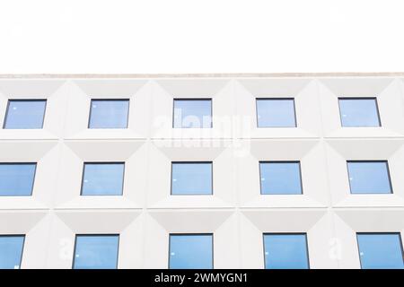 A minimalistic facade of a modern building with a geometric pattern of white structures and blue windows under a clear sky Stock Photo