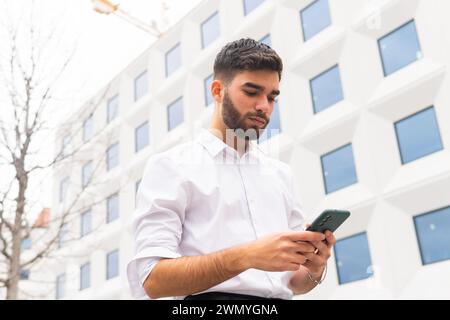 Young professional man focused on his smartphone with a modern geometric building facade in the background Stock Photo
