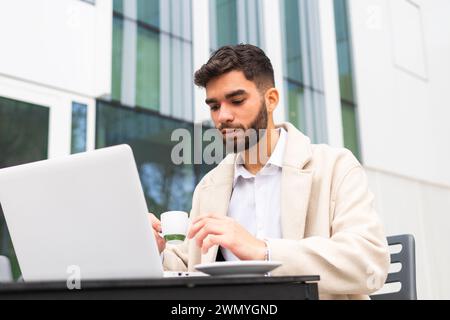 Concentrated businessman in a chic coat using a laptop and sipping coffee at an outdoor setting with a glass building backdrop Stock Photo