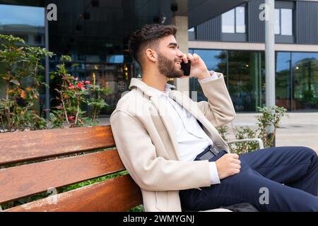 A relaxed businessman in a beige coat chats on his phone while sitting on a park bench, with an office building in the background Stock Photo