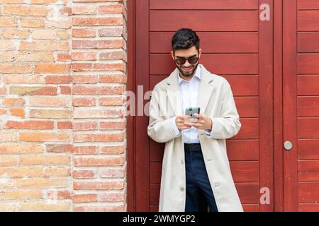 A joyful businessman in a beige coat and sunglasses using his phone, leaning against a brick wall next to a red door Stock Photo