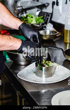 Close-up of a chef's hands with black gloves delicately plating a gourmet salad in a metal ring on a marble-patterned plate in a professional kitchen Stock Photo