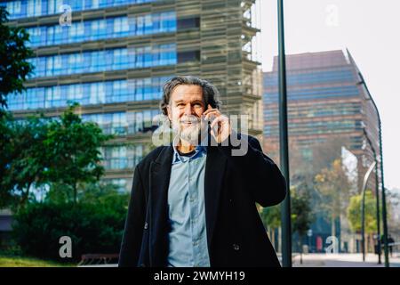 Smiling elderly man in a business suit engages in a phone conversation against an urban backdrop Stock Photo