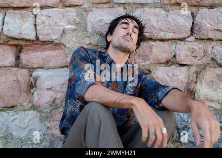 A young man with a pensive expression sits against an aged stone wall, appearing lost in thought Stock Photo