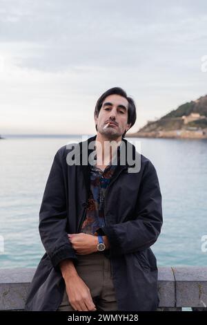 A pensive young man stands by the seaside, smoking a cigarette, dressed in a dark coat against a soft blue sky Stock Photo