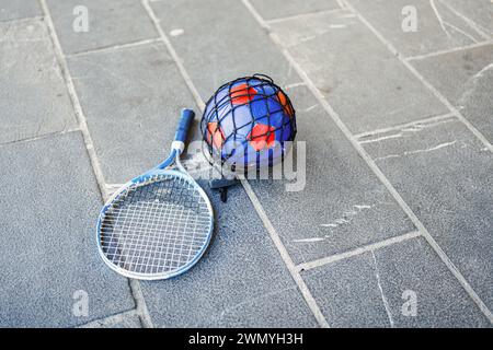 A badminton racket lying next to a soccer ball in a blue and red net on a concrete surface, symbolizing outdoor activities. Stock Photo