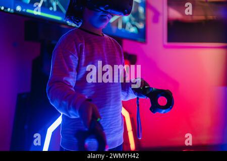 Child focused on playing with a VR headset and controllers in a room illuminated by neon gaming lights Stock Photo