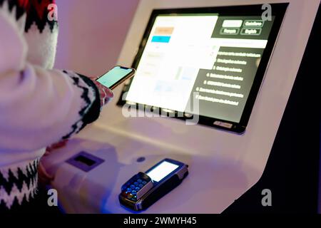 Anonymous person using a smartphone to interact with a digital kiosk touch screen display next to a card payment terminal Stock Photo
