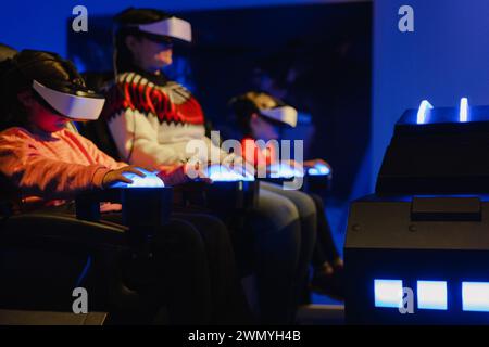 Row of focused individuals using VR headsets and hand controls in high-tech simulation seats with blue lighting Stock Photo