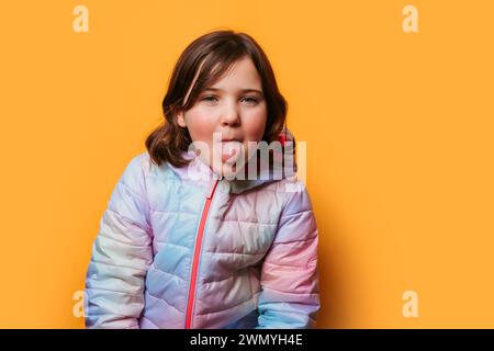 Playful girl sticking out her tongue, dressed in a multicolored jacket against a vibrant orange background Stock Photo
