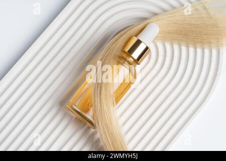 A luxurious hair care oil in a sleek glass bottle placed alongside a lock of healthy blonde hair, perfectly staged on a white corrugated surface. Stock Photo