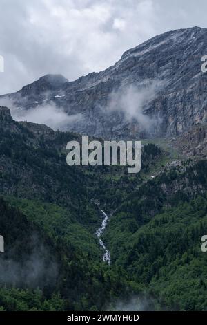 Majestic view of cloud-covered peaks in the Bujaruelo Valley, with lush green foliage and cascading waterfall visible in the middle distance. Stock Photo