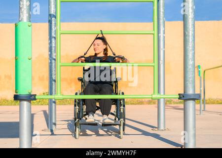 A focused woman in a wheelchair uses resistance bands for an upper body workout at an outdoor gym Stock Photo