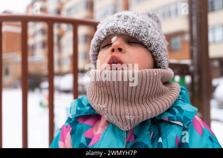 Child in a colorful jacket catching snowflakes with mouth open against a playground backdrop Stock Photo