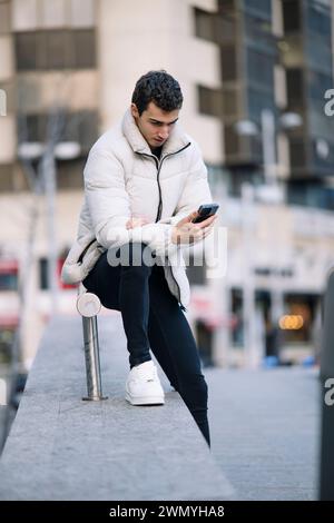 A young man in a white jacket is focused on his smartphone while sitting on a metal post in an urban setting Stock Photo