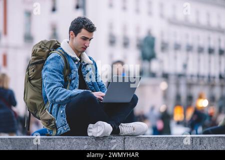 Young man works on his laptop while seated on the steps in a lively Madrid square Stock Photo