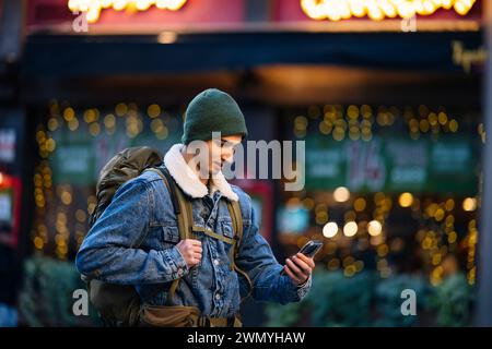 Young man in a beanie and denim jacket using his phone, with twinkling city lights in the background Stock Photo