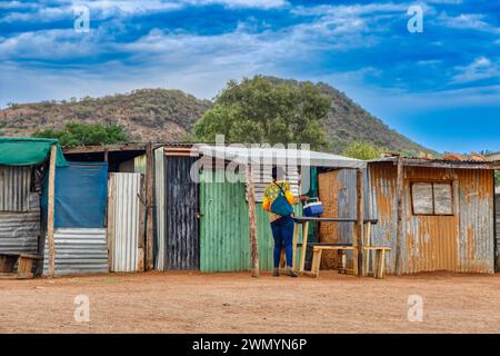 township informal settlement in africa near a hill, woman caring a cooler box in front of the corrugated iron shacks Stock Photo