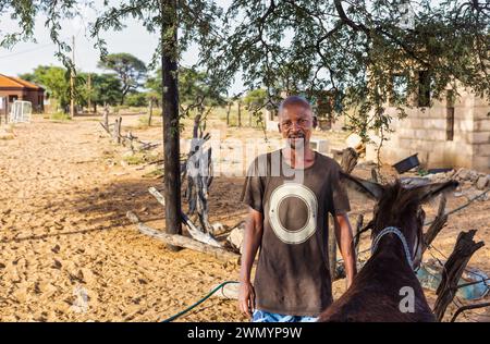 african village man on a dirt road holding his donkey, houses from the village in the background Stock Photo