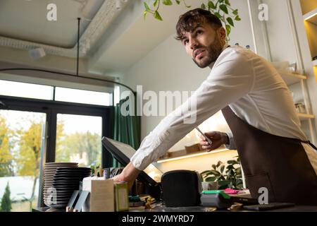Male barista at counter using cashbox computer in cafe store checking client's order Stock Photo