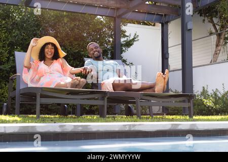 African American man and biracial woman relax on poolside loungers at home, sharing a joyful moment Stock Photo