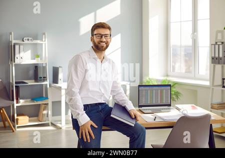 Young business man sitting on the desk of his workplace at office, looking at camera and smiling. Stock Photo