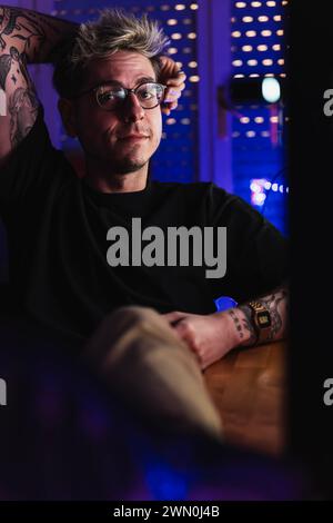 Young tattooed gamer wearing glasses sitting on the chair of his room in his leisure time Stock Photo