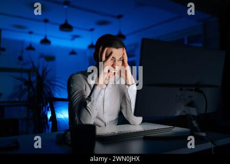 Overworked stressed young man working office at night on computer. Excited tired worker male angry and mad screaming frustrated and furious, shouting Stock Photo