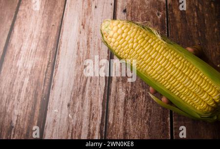 A close-up photo of several fresh corn cobs with vibrant green husks, arranged on a weathered wooden surface Stock Photo
