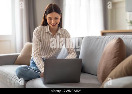 Happy young freelancer woman using laptop on comfortable sofa Stock Photo