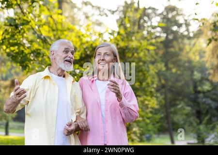 Joyful elderly couple engaging in a heartwarming talk during a leisurely stroll in a verdant park, basking in golden sunlight. Stock Photo