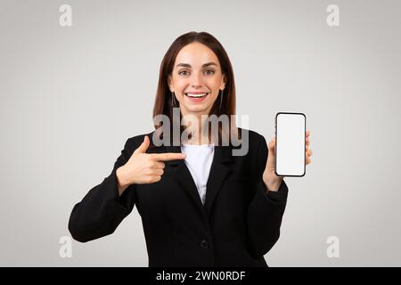 Enthusiastic businesswoman in a sleek black suit happily presenting a smartphone with a blank screen Stock Photo