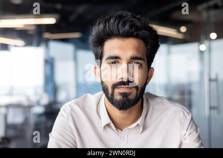 Close-up photo of a serious young Indian man with a beard and a shirt working in the office, sitting and looking confidently into the camera. Stock Photo