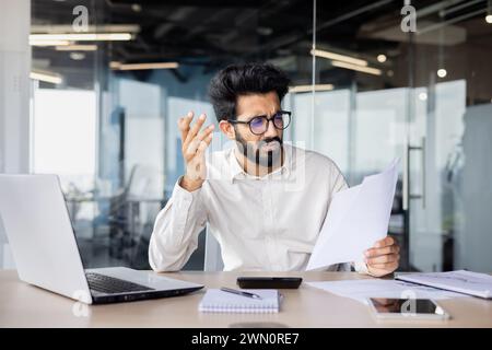 Frustrated young indian male businessman working in office at desk with documents, looking disappointed at papers and bills. Stock Photo