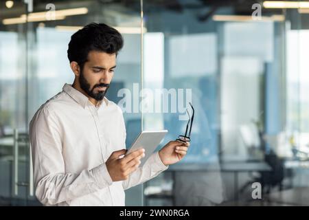 Indian young male programmer standing in the office center indoors and looking focused at the screen of the tablet he is holding in his hands and wearing glasses. Stock Photo