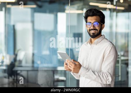 Portrait of a successful Indian young male programmer engineer wearing glasses standing inside the office, holding a tablet and smiling at the camera. Stock Photo