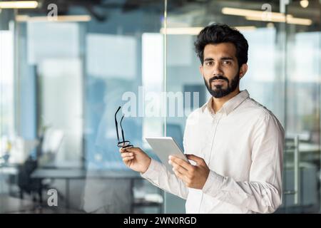 Portrait of a young Indian male programmer, developer, specialist standing in the office, holding glasses and a tablet, confidently looking at the camera. Stock Photo