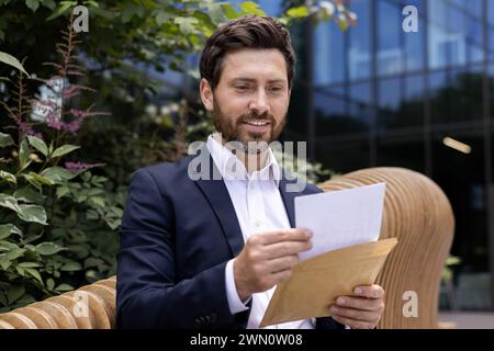 Cheerful male professional in a suit reviews a manila envelope contents outside a modern building with foliage. Stock Photo