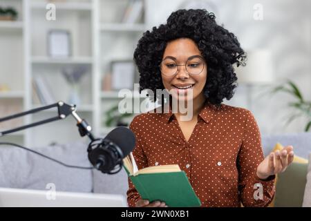 Home recording online studio. Young smiling African American woman sitting on sofa in front of microphone and laptop and reading book, falling asleep to podcast, giving sermon. Stock Photo