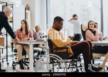 Handicapped male employee in a wheelchair seamlessly collaborates with colleagues, searching for information on his phone during work. Stock Photo