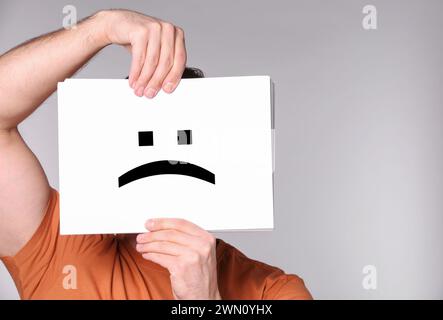 Man hiding behind sheet of paper with sad face on grey background Stock Photo