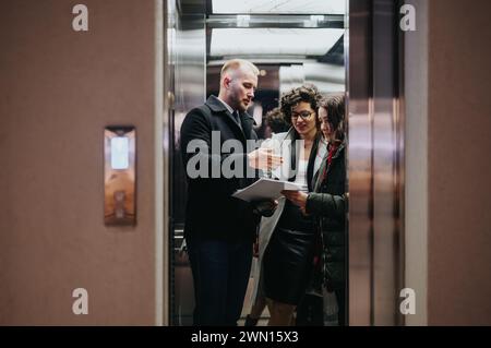 Two women and one man in professional attire engage in a discussion while holding papers and a digital tablet in an office elevator corridor. Stock Photo