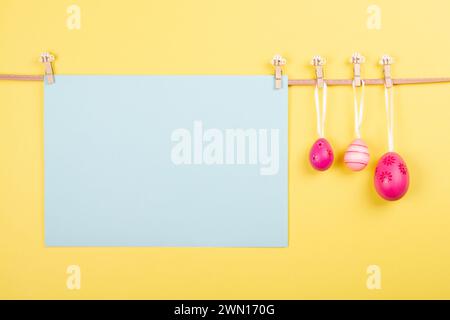 Blank light blue sheet of paper and three pink Easter eggs hang at rope on pastel yellow background. Easter card with open area for your words or imag Stock Photo