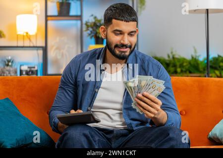 Planning budget. Rich happy Indian man using calculator counting money cash, calculate domestic bills at home. Hispanic guy satisfied of income earnings win saves money for planned vacation gifts. Stock Photo